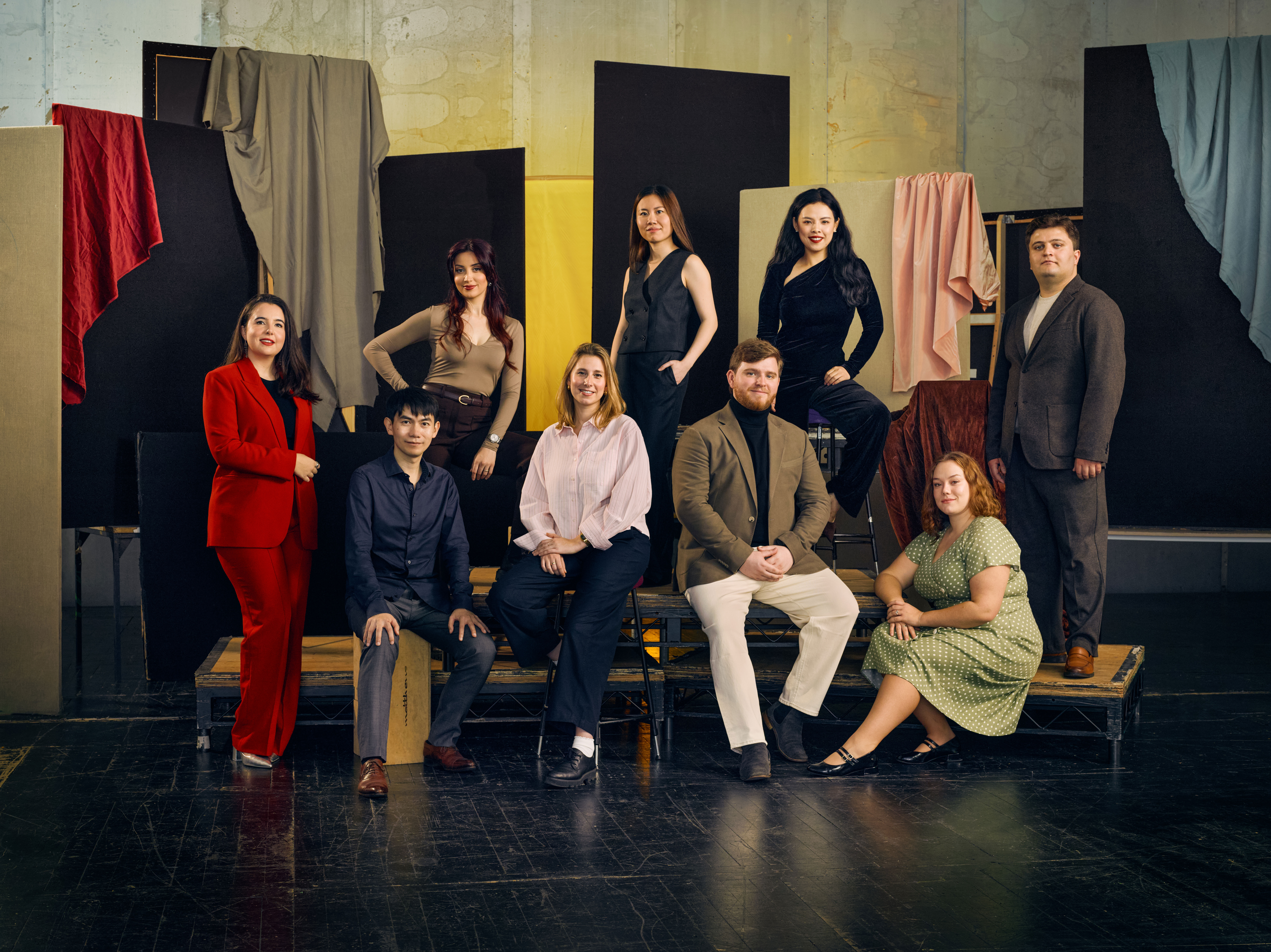 A group of nine people in formal attire pose against a backdrop of hanging fabrics in various colours. 