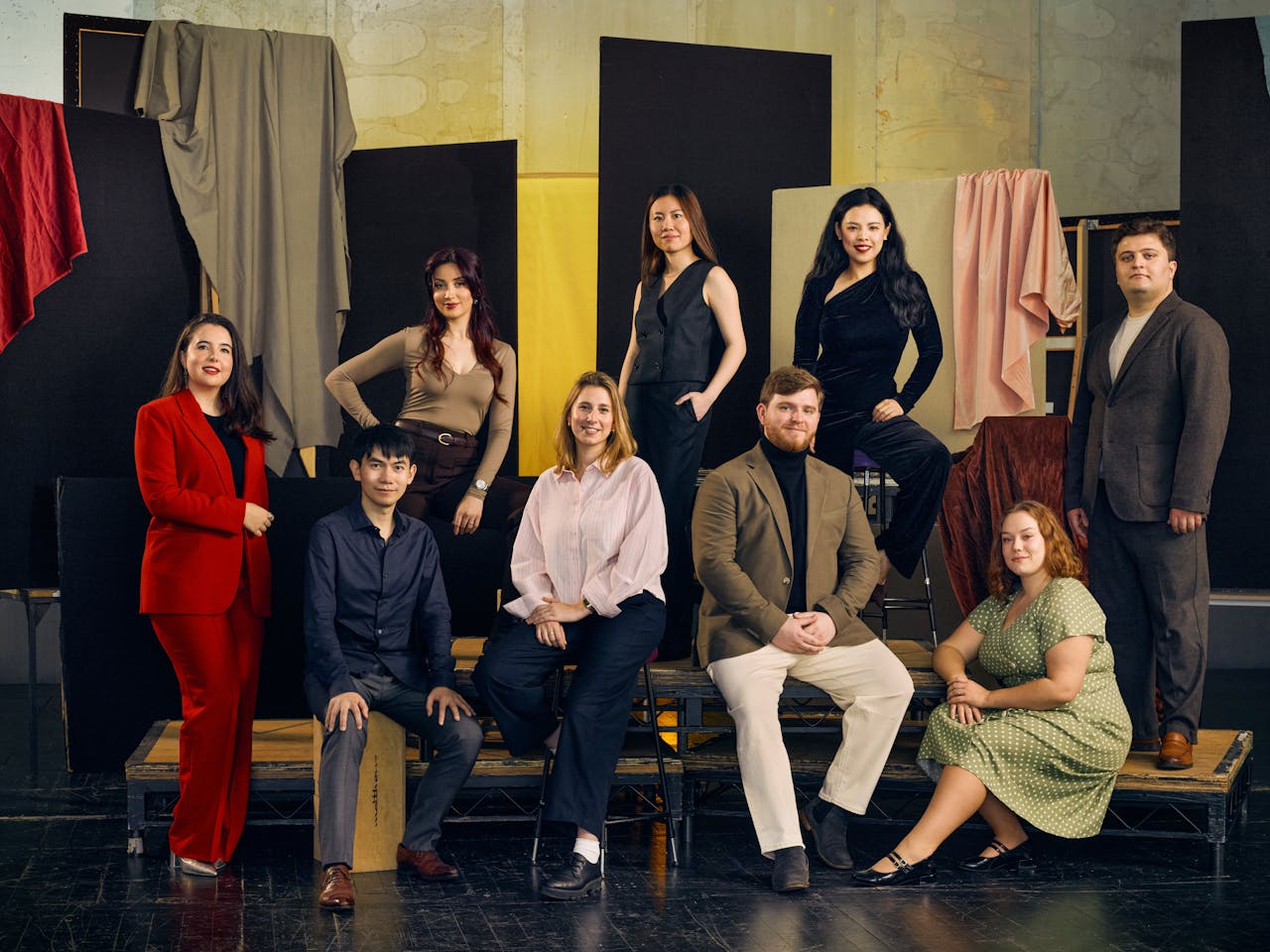 A group of nine people in formal attire pose against a backdrop of hanging fabrics in various colours.