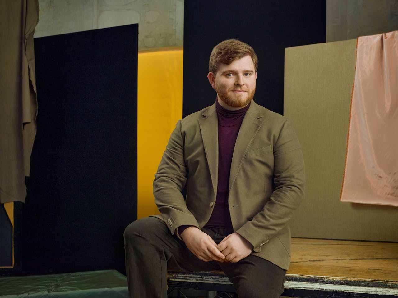 Jette Parker Artist Sam Hird sits in the centre of the photo, he is wearing a burgundy turtle neck jumper and brown suit jacket and trousers.