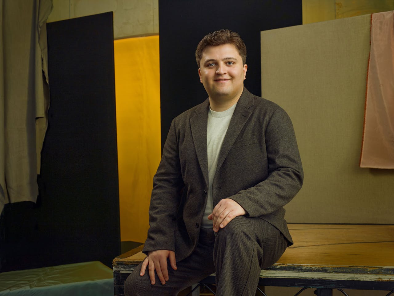 Jette Parker Artist Giorgi Guliashvili sits in the centre of the photo, he is wearing a white t-shirt and brown tweed suit jacket and trousers.