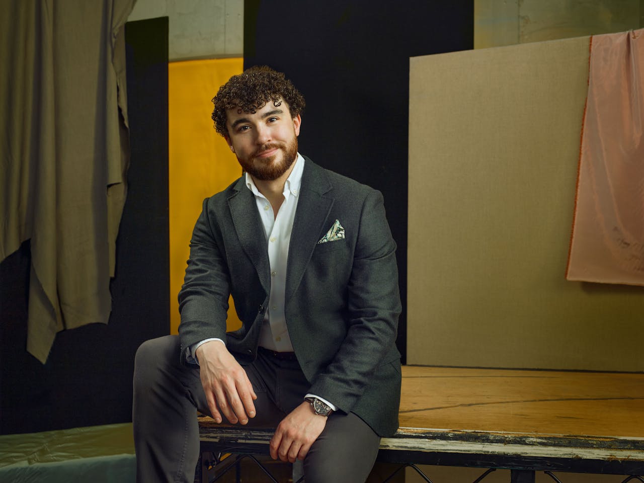 Jette Parker Artist Ossian Huskinson sits in the centre of the photo, he is wearing a white shirt unbuttoned at the collar and charcol grey suit jacket and trousers. He has a green silk pocket square in his jacket breast pocket.