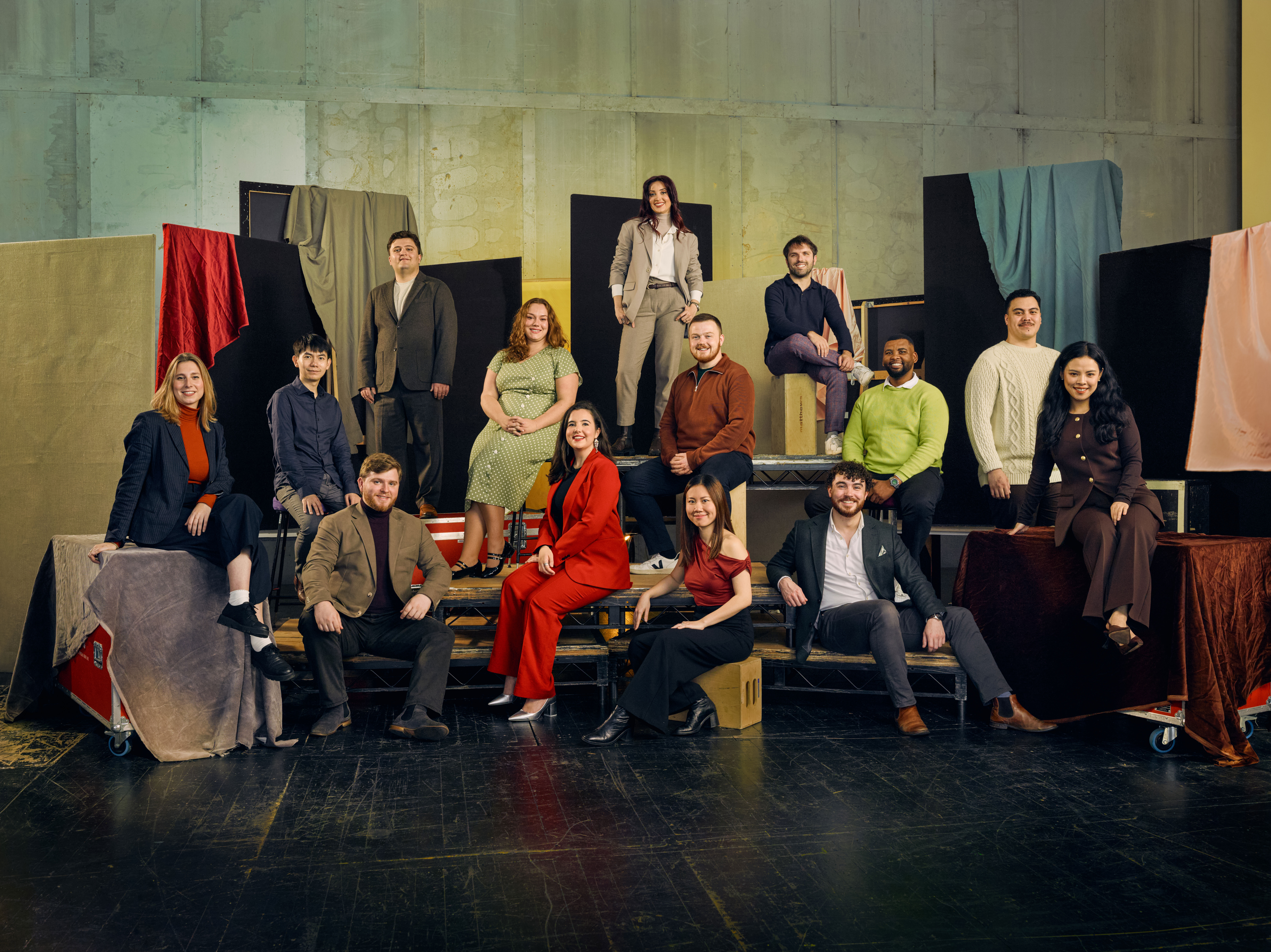 A group of 13 people pose in colourful formal clothing against a textured backdrop with fabric drapes. They are seated on various platforms. 