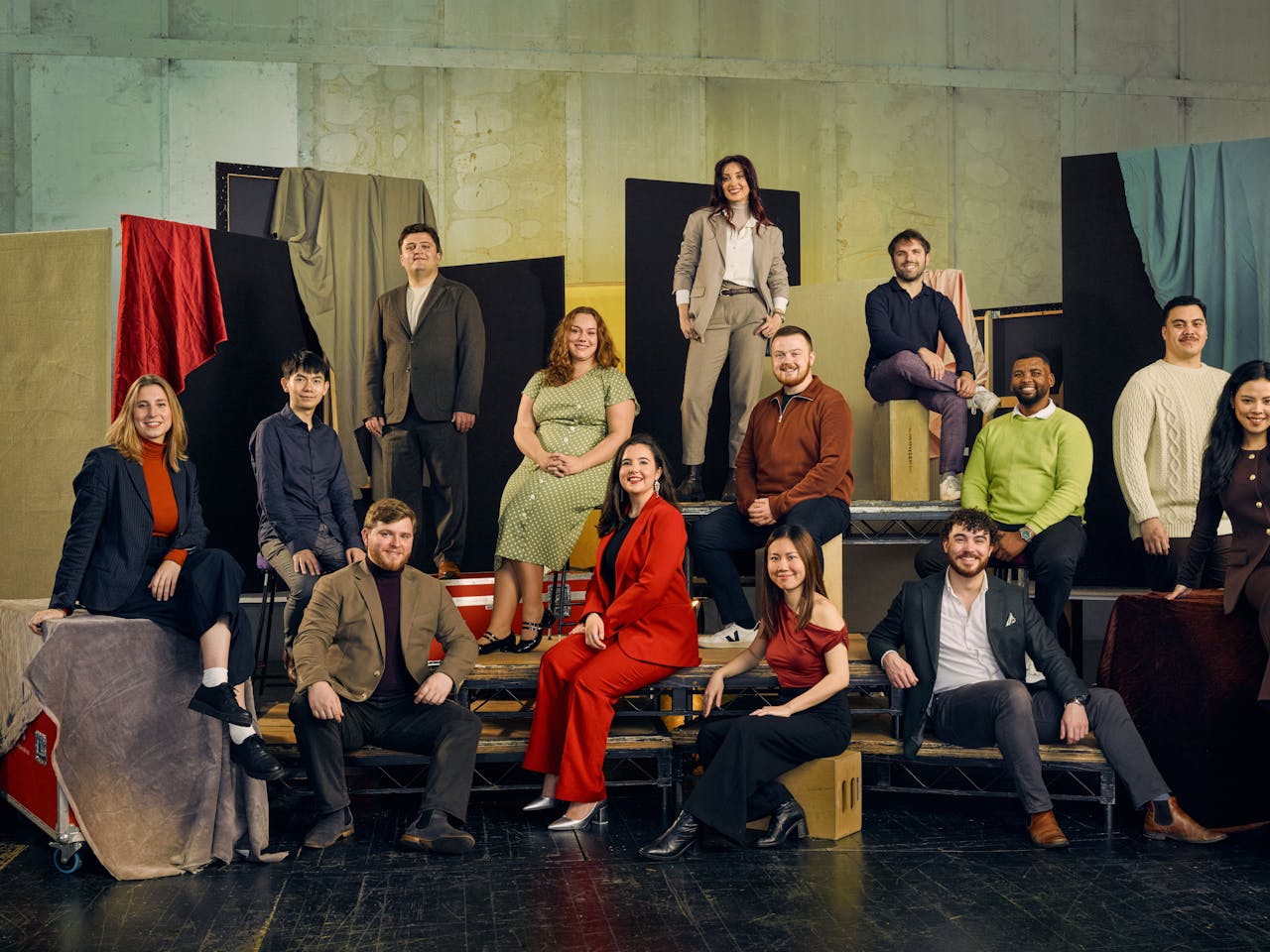 A group of 13 people pose in colourful formal clothing against a textured backdrop with fabric drapes. They are seated on various platforms.