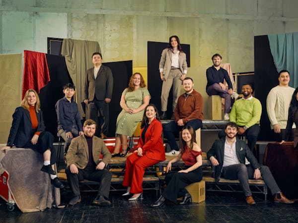 A group of 13 people pose in colourful formal clothing against a textured backdrop with fabric drapes. They are seated on various platforms.