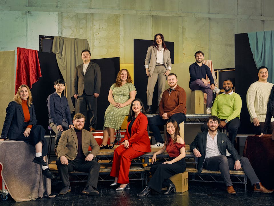 A group of 13 people pose in colourful formal clothing against a textured backdrop with fabric drapes. They are seated on various platforms.