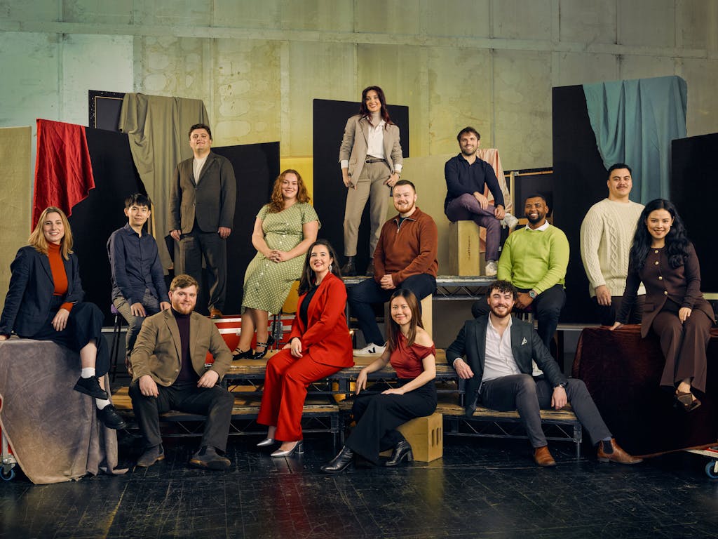 A group of 13 people pose in colourful formal clothing against a textured backdrop with fabric drapes. They are seated on various platforms.