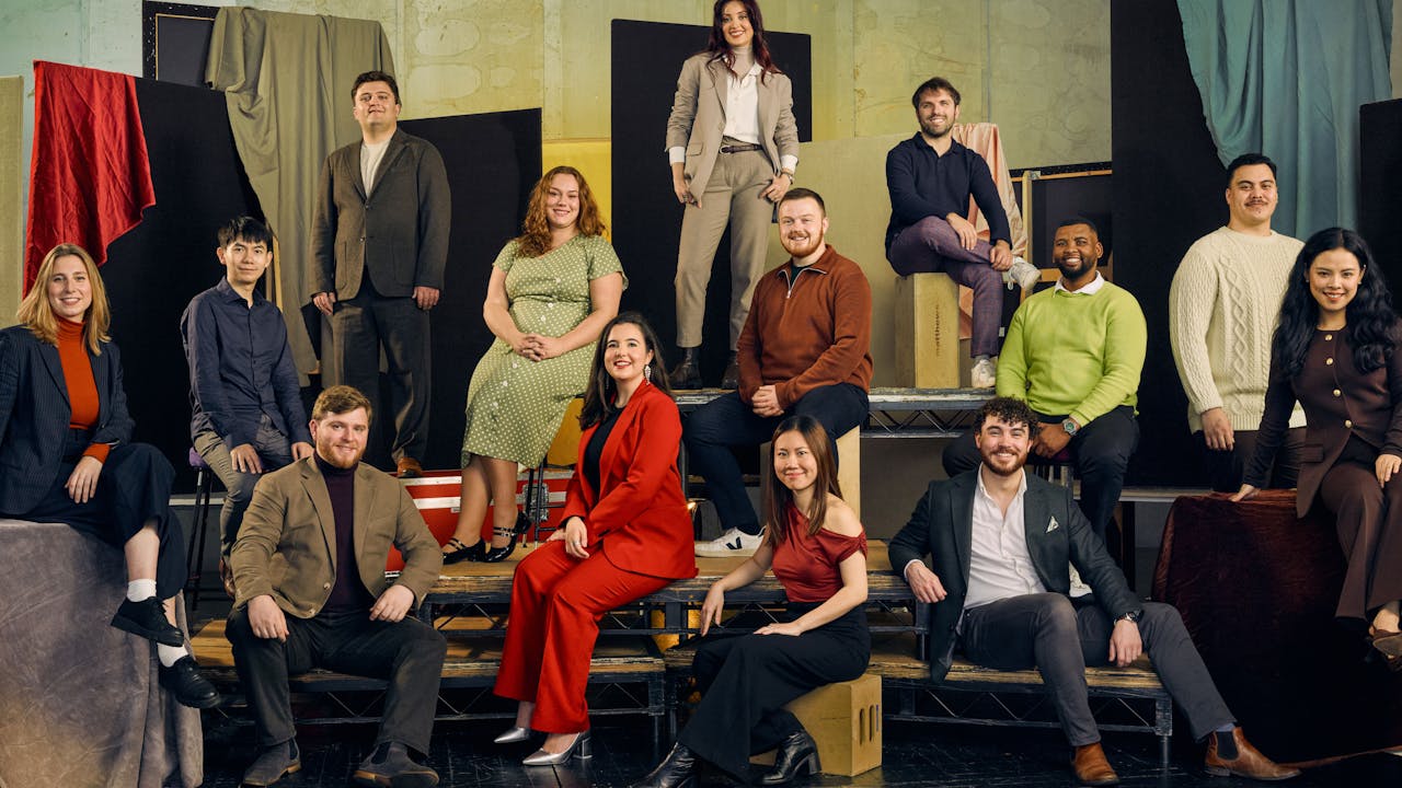 A group of 13 people pose in colourful formal clothing against a textured backdrop with fabric drapes. They are seated on various platforms.