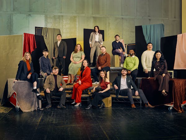 A group of 13 people pose in colourful formal clothing against a textured backdrop with fabric drapes. They are seated on various platforms.
