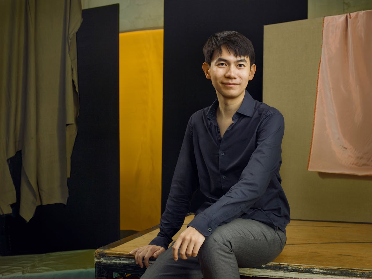 Jette Parker Artist Richard Fu sits in the centre of the photo, he is wearing a navy shirt unbuttoned at the collar and grey suit trousers.