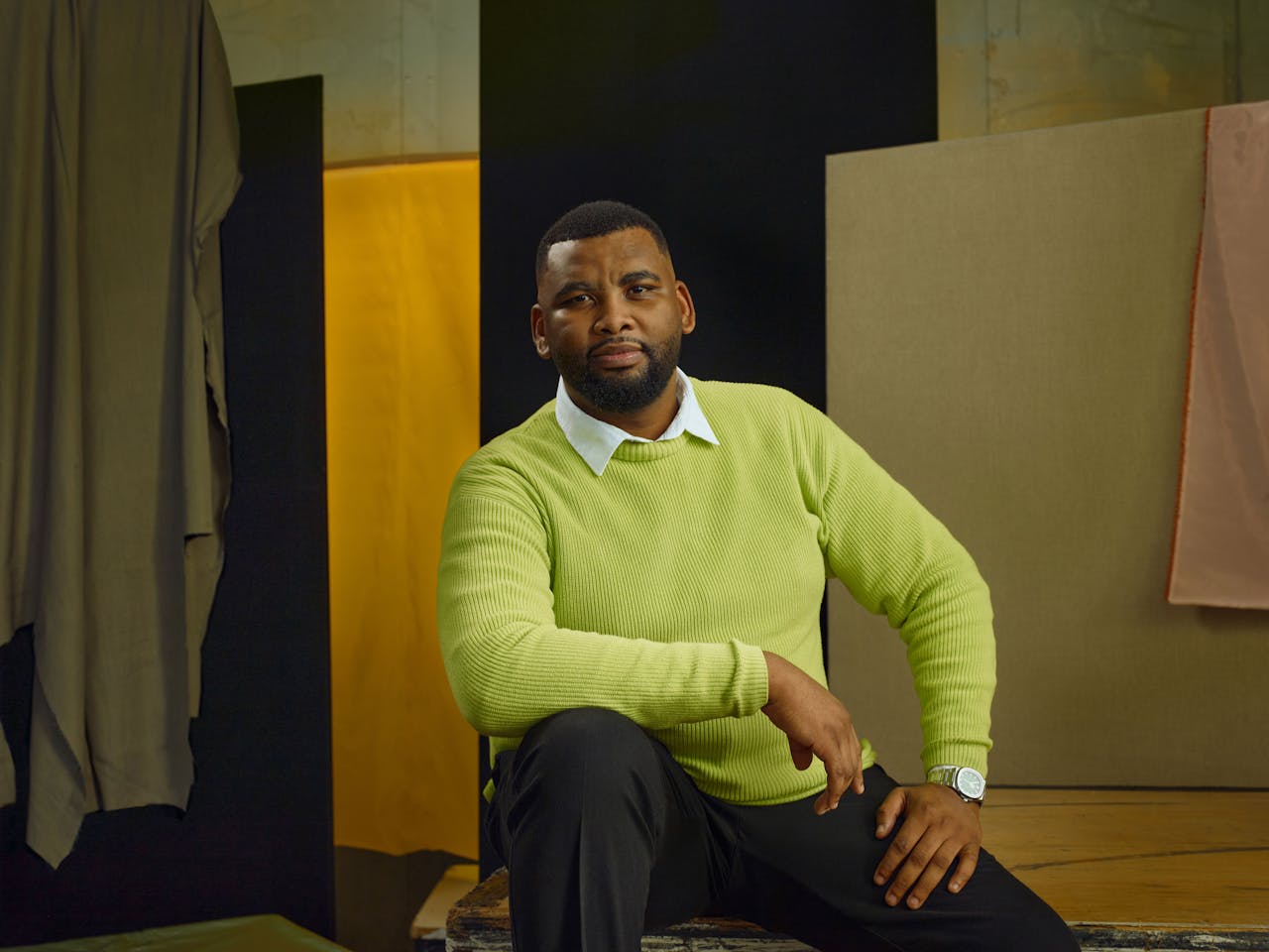 Jette Parker Artist Siphe Kwani sits in the centre of the photo, he is wearing a lime green jumper over a white collared shirt and black suit trousers.