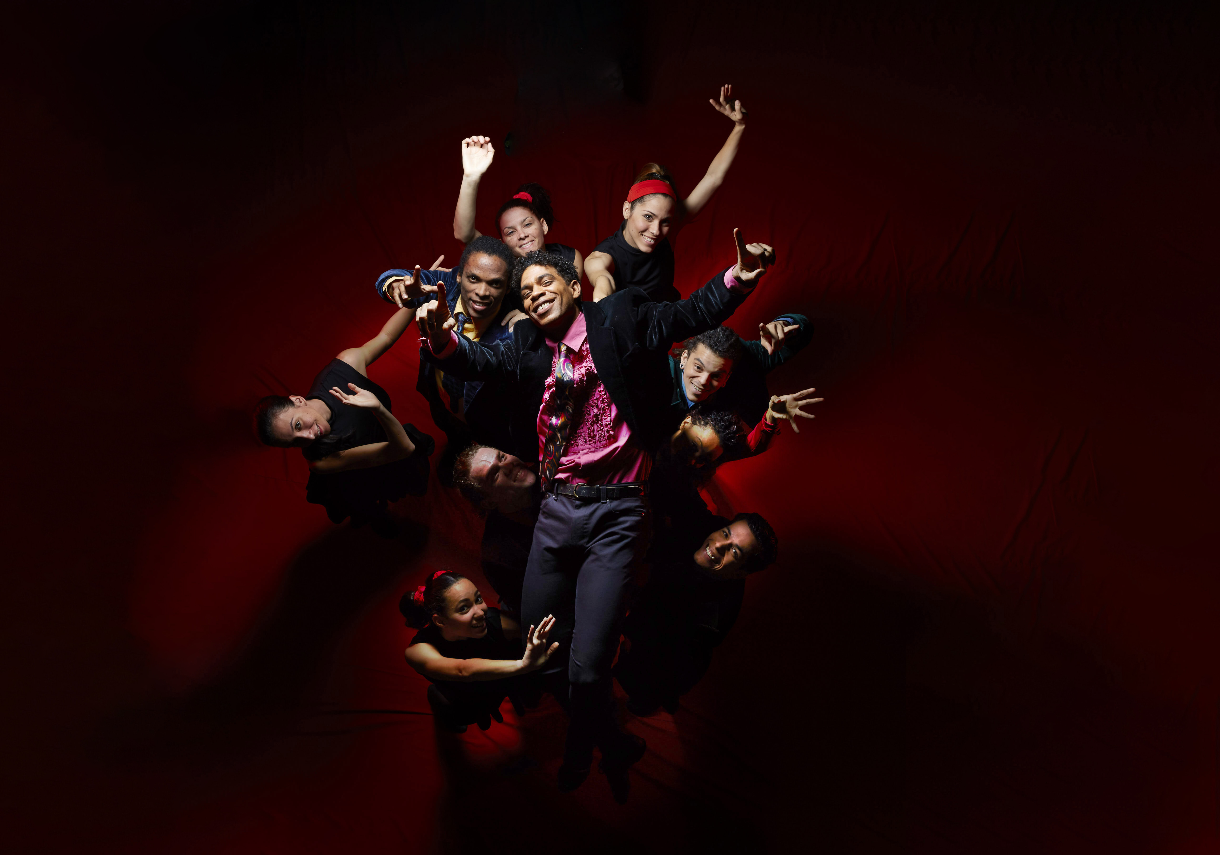 Ballet Dancer Carlos Acosta stands in front of a group of people. He wears a pink shirt and black jacket. Everyone's arms are outstretched.