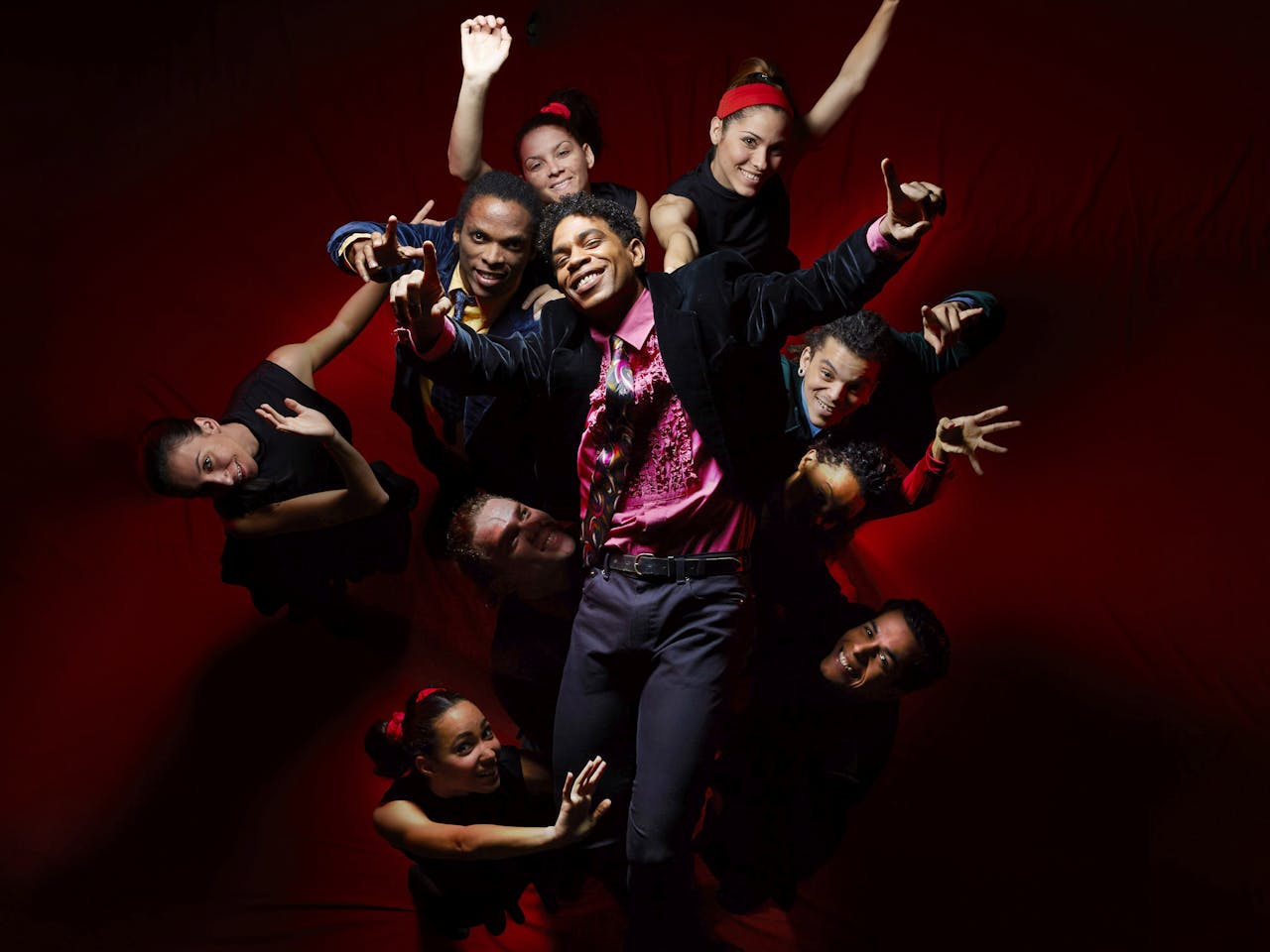 Ballet Dancer Carlos Acosta stands in front of a group of people. He wears a pink shirt and black jacket. Everyone's arms are outstretched.
