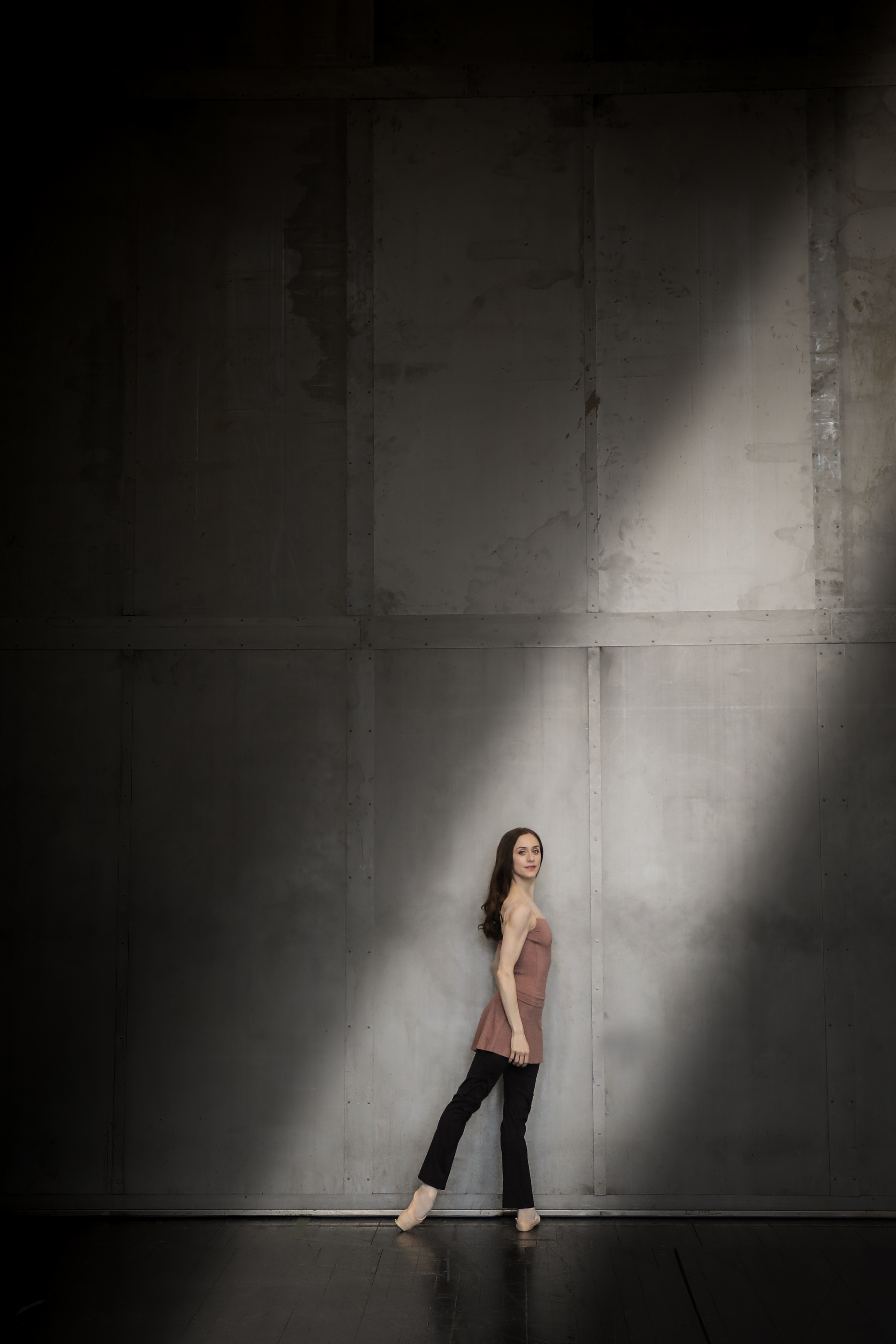 A dancer stands against a grey wall in a shaft of light. She has long, straight dark hair and is wearing a pink top, black trousers and pointe shoes.