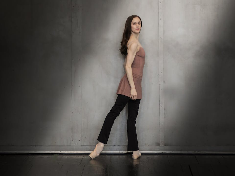 A dancer stands against a grey wall in a shaft of light. She has long, straight dark hair and is wearing a pink top, black trousers and pointe shoes.