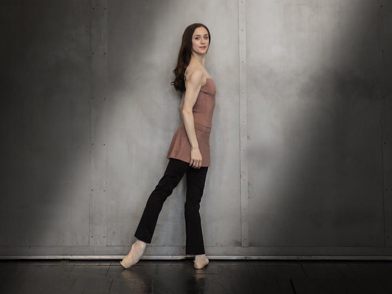 A dancer stands against a grey wall in a shaft of light. She has long, straight dark hair and is wearing a pink top, black trousers and pointe shoes.