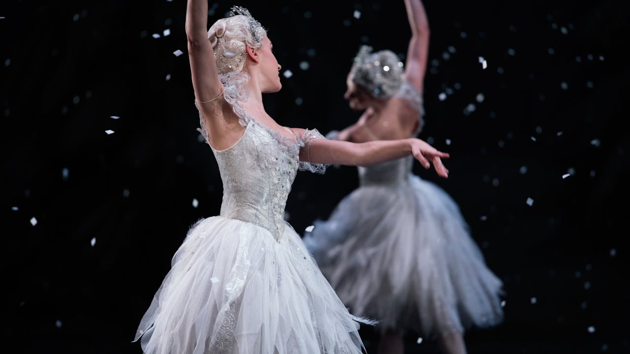 Two dancers, dressed in white, pose near each other. They dance against a black background with white snow falling from above.