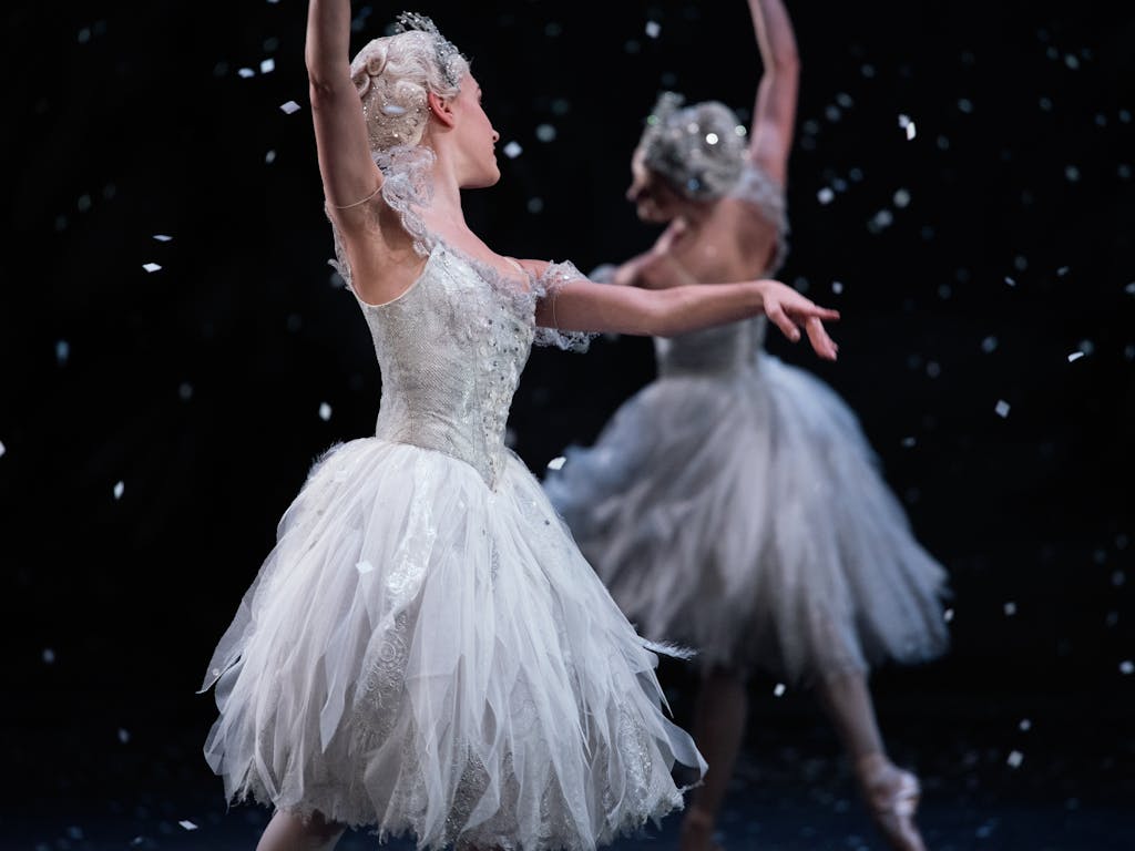Two dancers, dressed in white, pose near each other. They dance against a black background with white snow falling from above.