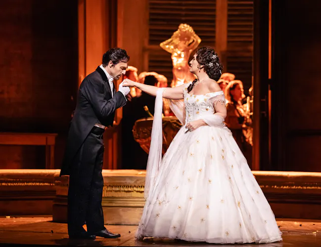 Violetta stands centre stage in a long white ball gown, Alfredo holds her outstretched hand and kisses. Alfredo wears a dinner suit - they stand in front of an ornate fountain.