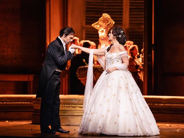 Violetta stands centre stage in a long white ball gown, Alfredo holds her outstretched hand and kisses. Alfredo wears a dinner suit - they stand in front of an ornate fountain.