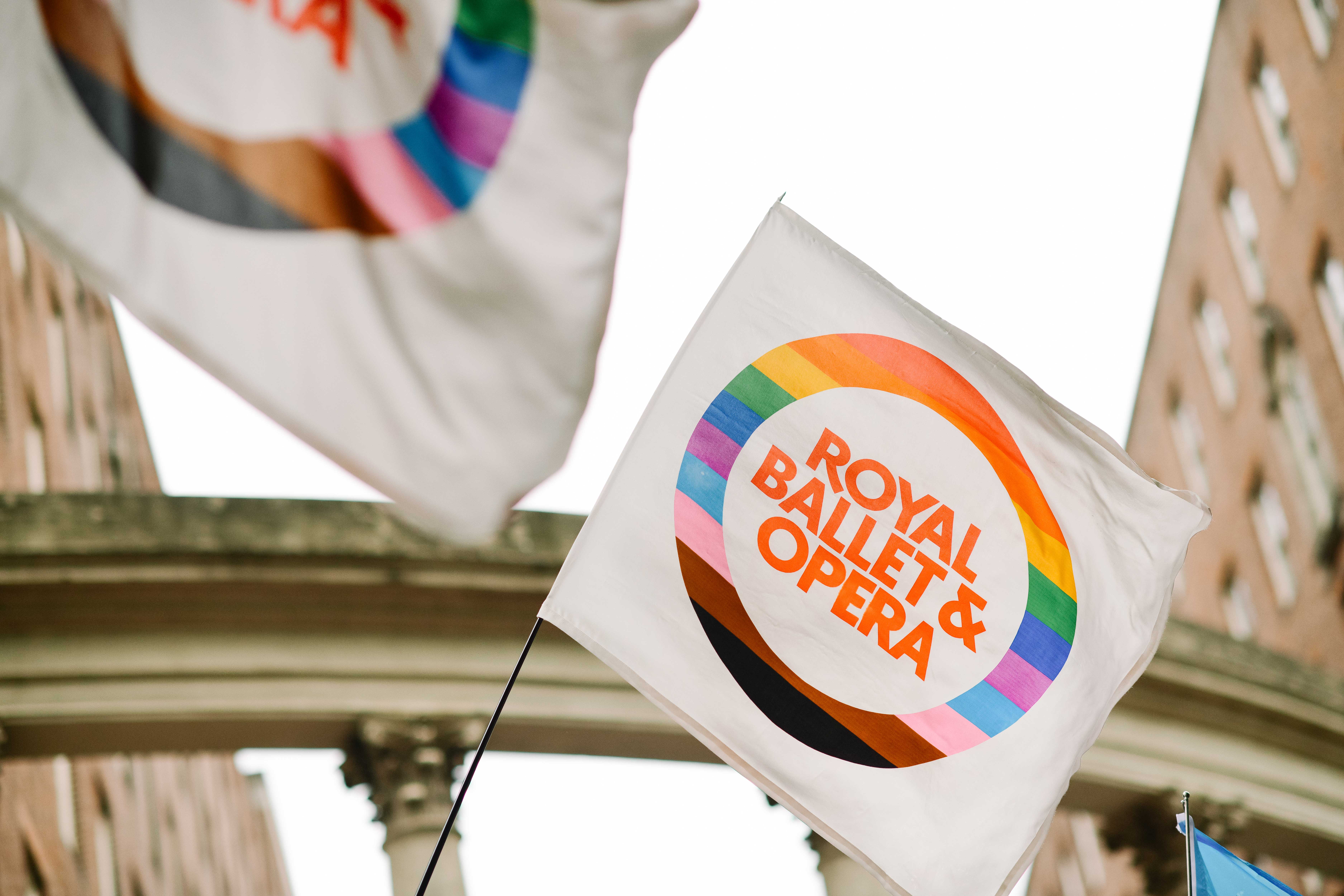 A white flag with a vibrant rainbow border reads "Royal Ballet & Opera" in bold orange, waving against a backdrop of historic brick buildings and an archway.
