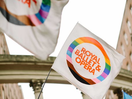 A white flag with a vibrant rainbow border reads "Royal Ballet & Opera" in bold orange, waving against a backdrop of historic brick buildings and an archway.