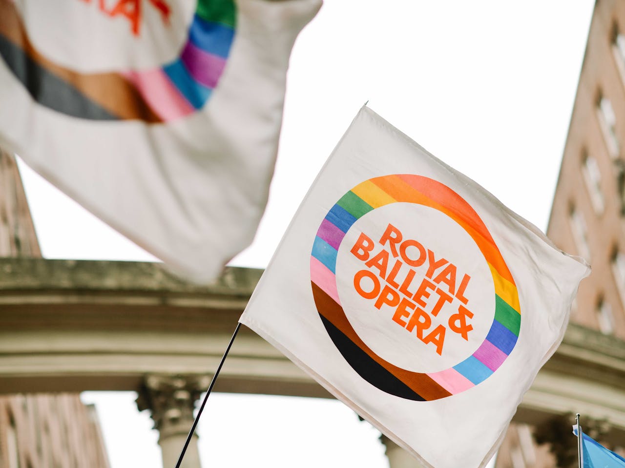 A white flag with a vibrant rainbow border reads "Royal Ballet & Opera" in bold orange, waving against a backdrop of historic brick buildings and an archway.