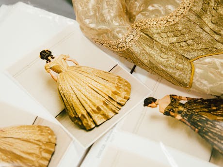 Gold brocade fabric lies on a table next to costume photos cards featuring actors in 1830's style dresses.