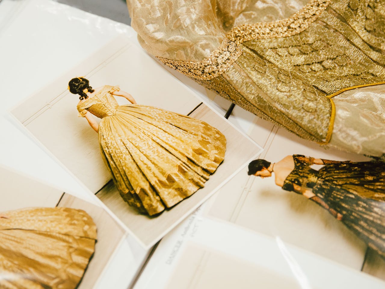 Gold brocade fabric lies on a table next to costume photos cards featuring actors in 1830's style dresses.
