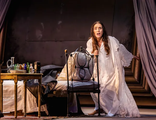 A distressed woman in a white gown holds on to a iron chair.