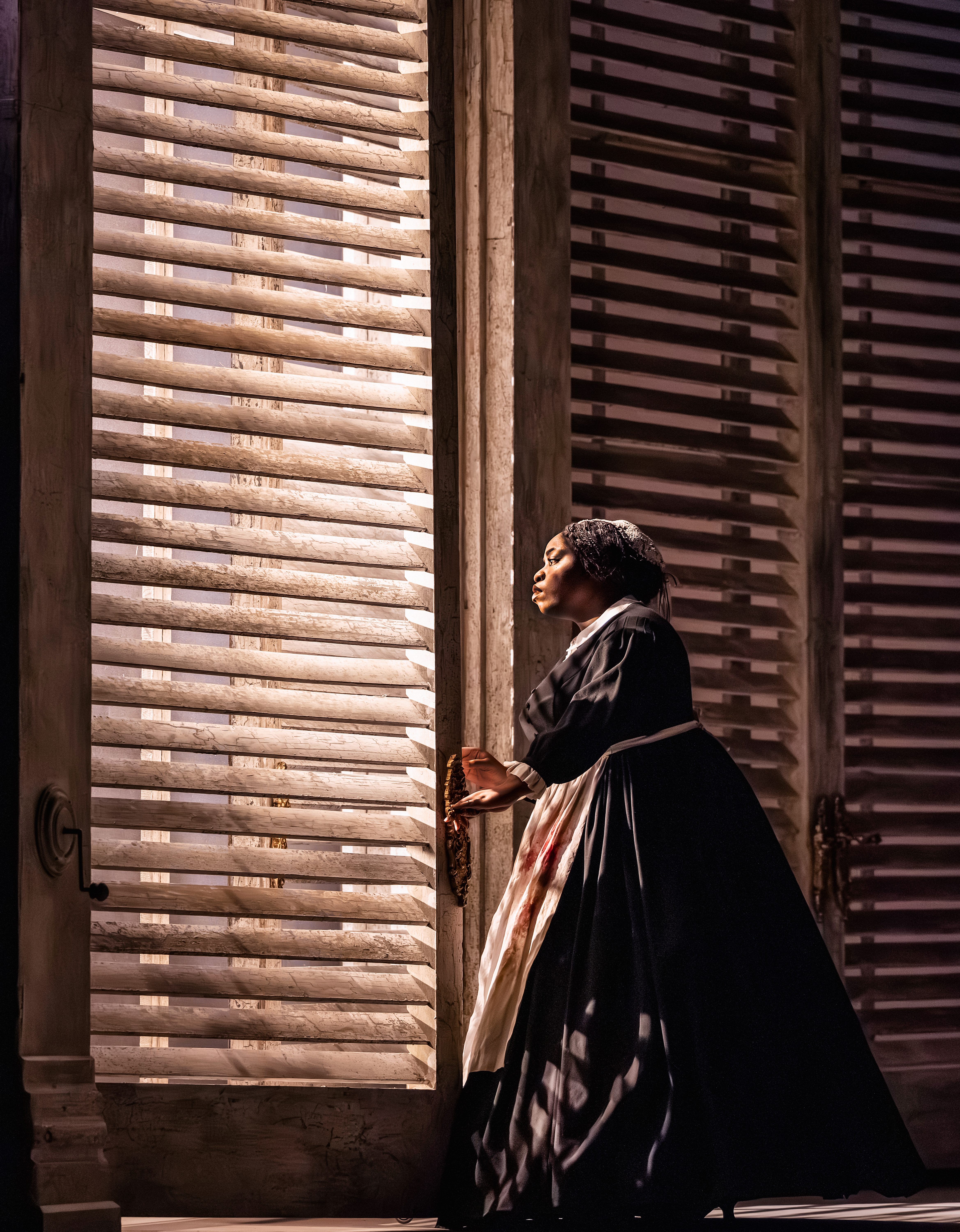 A woman in a black dress looks out through a slatted window.