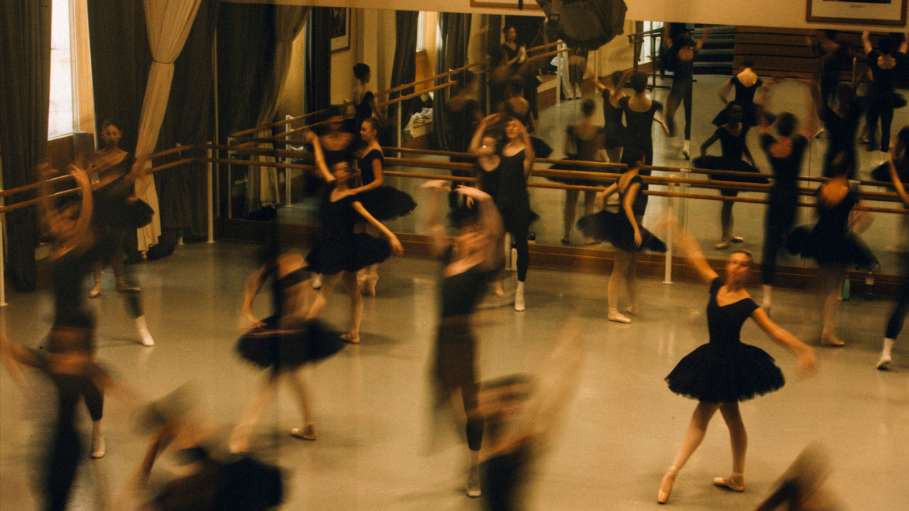 Members of The Royal Balelt School rehearse in a studio. They are dressed in black and hold various positions. Some of the dancers are blurred as they move.