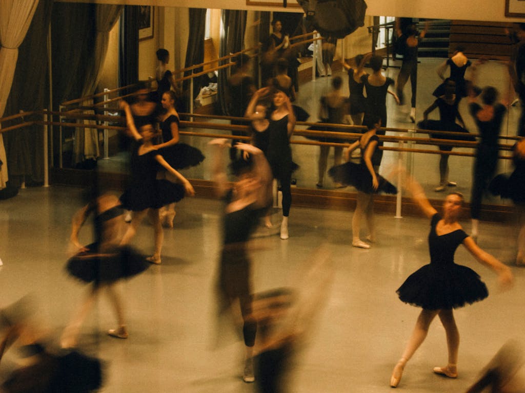 Members of The Royal Balelt School rehearse in a studio. They are dressed in black and hold various positions. Some of the dancers are blurred as they move.