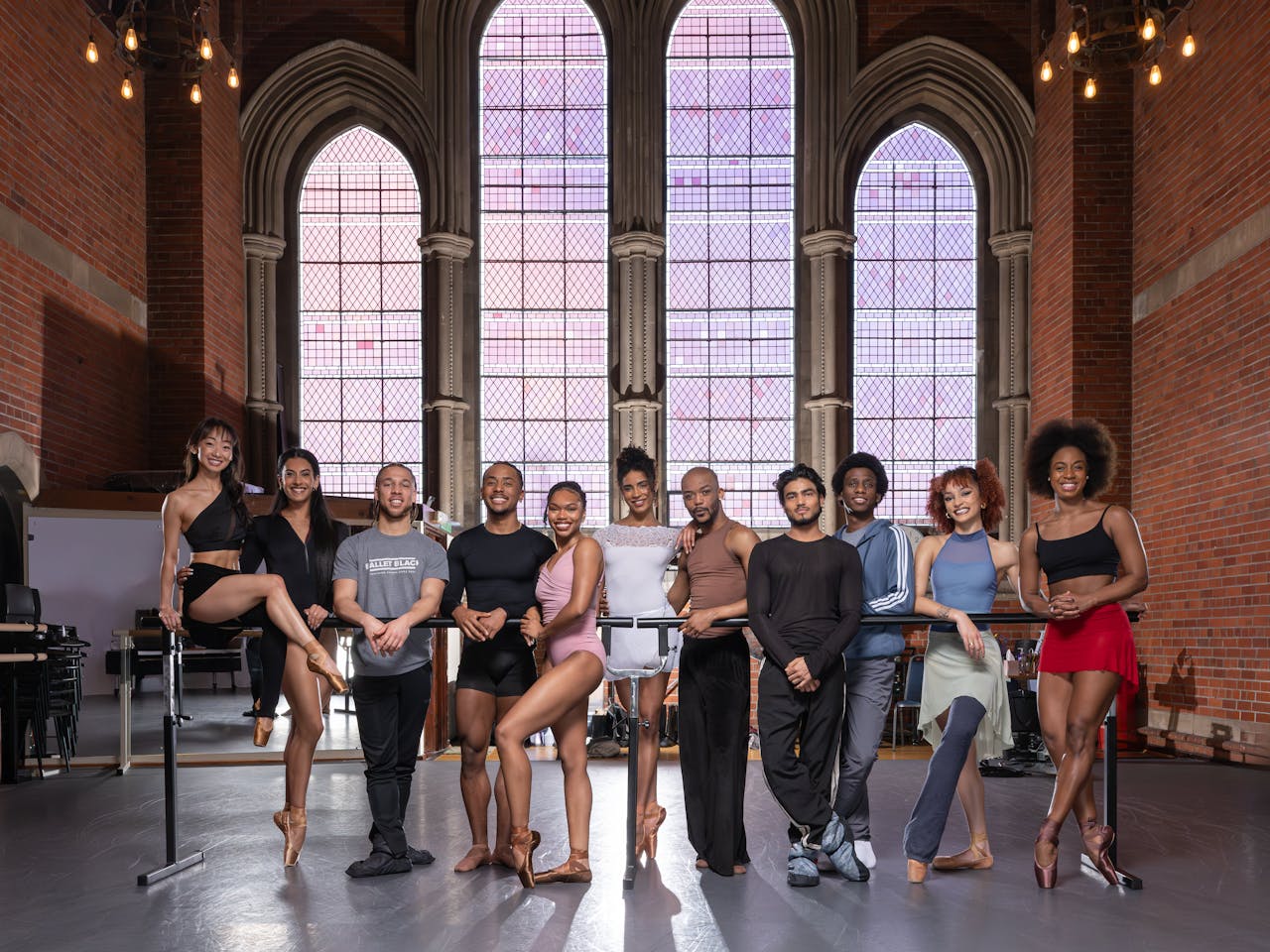 A group of people in sportswear lean on a ballet barre in the centre of a room. Behind them are large stained glass windows.