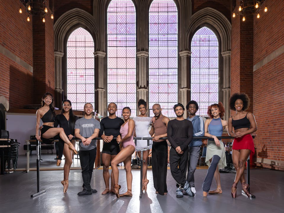 A group of people in sportswear lean on a ballet barre in the centre of a room. Behind them are large stained glass windows.