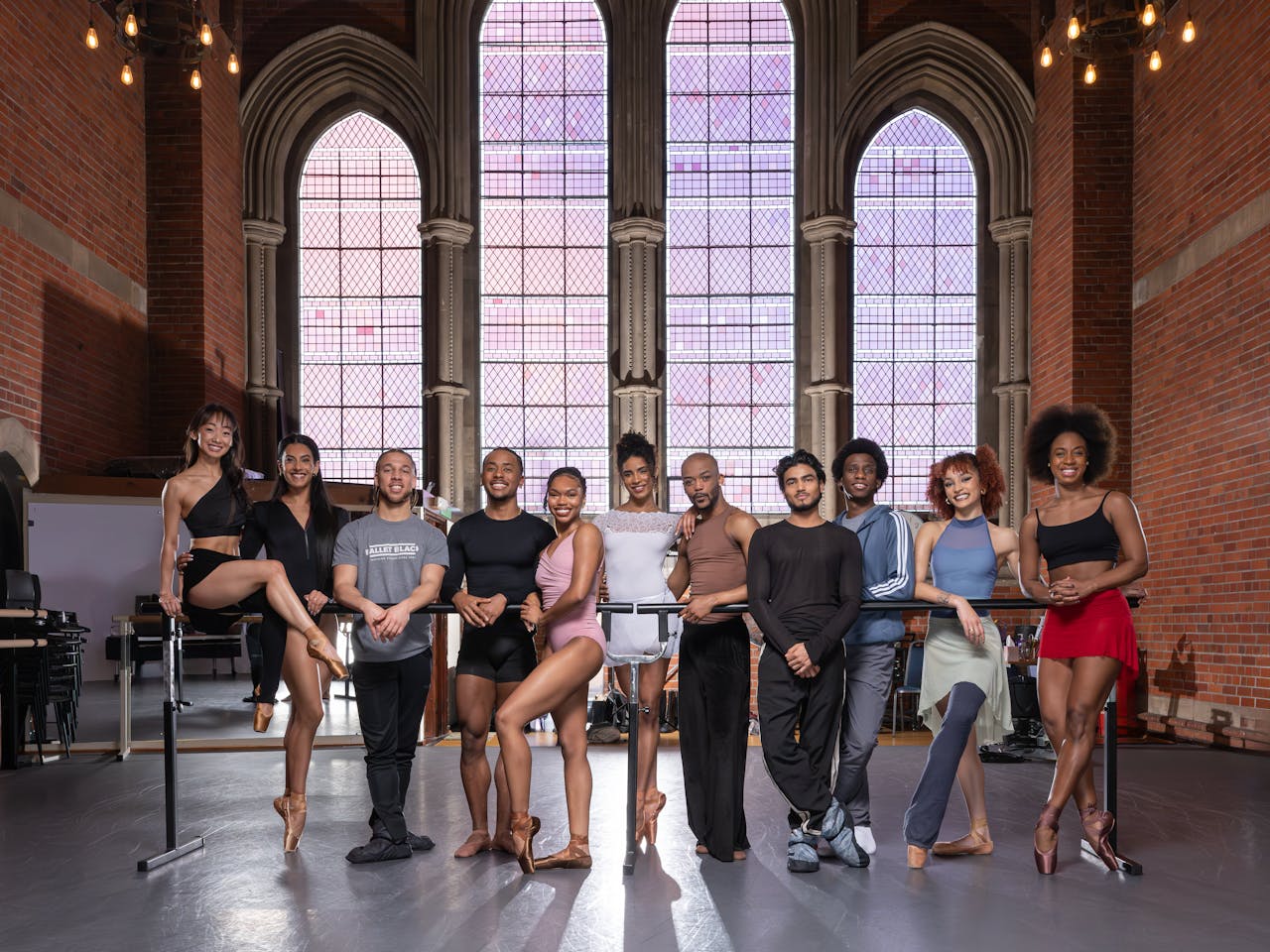 A group of people in sportswear lean on a ballet barre in the centre of a room. Behind them are large stained glass windows.