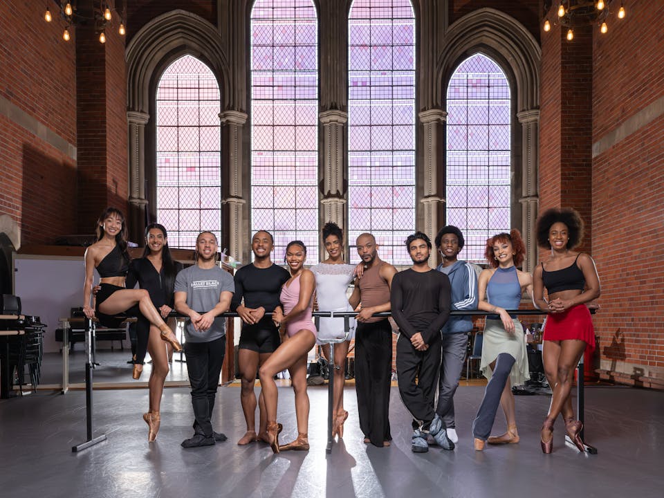 A group of people in sportswear lean on a ballet barre in the centre of a room. Behind them are large stained glass windows.