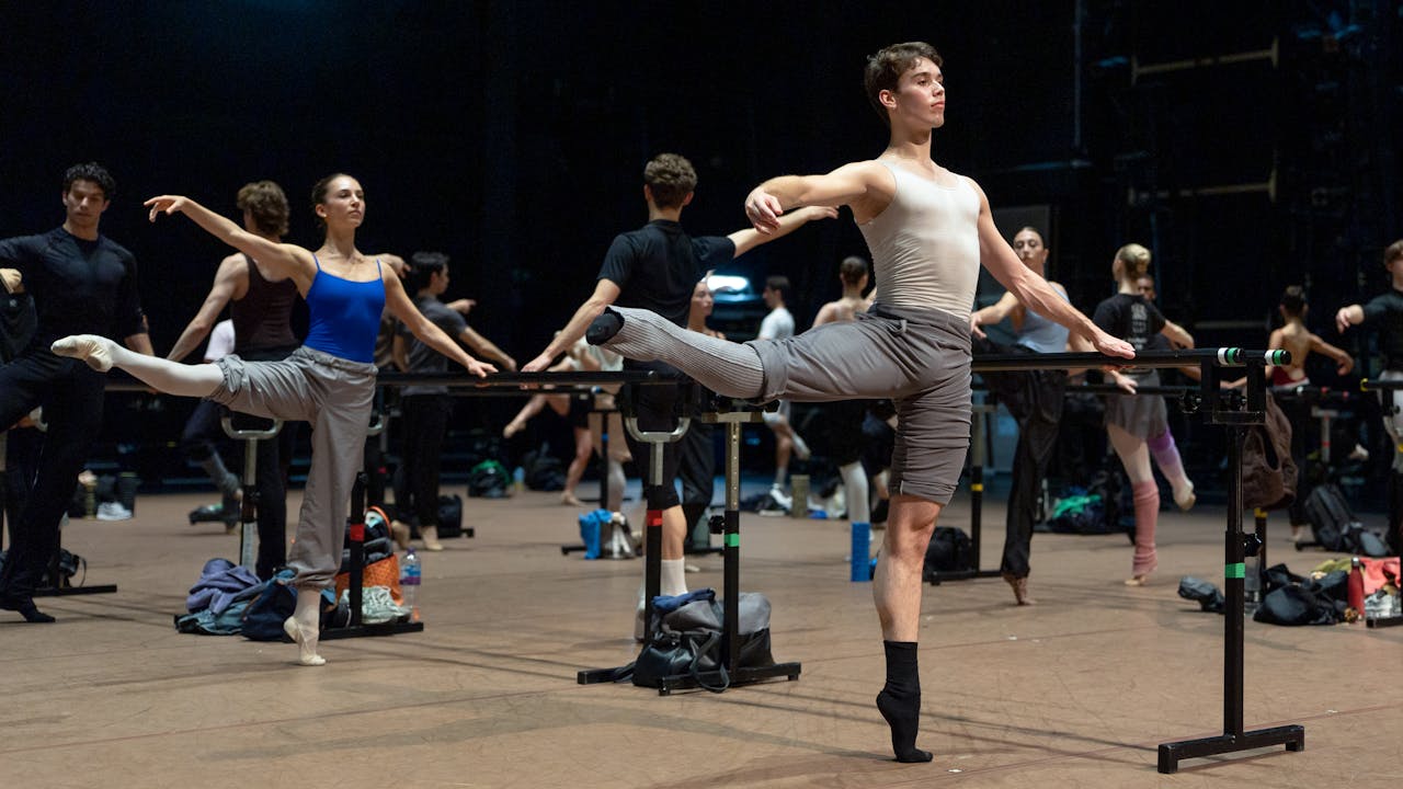 Dancers warm up at the barre. A man in grey trousers and a white t-shirt balances on one leg. Behind him is a women in a blue leotard and grey trousers, also balances on one leg.