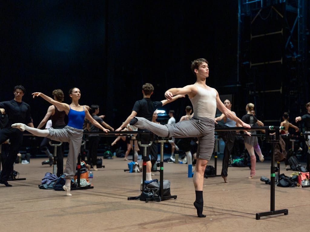 Dancers warm up at the barre. A man in grey trousers and a white t-shirt balances on one leg. Behind him is a women in a blue leotard and grey trousers, also balances on one leg.