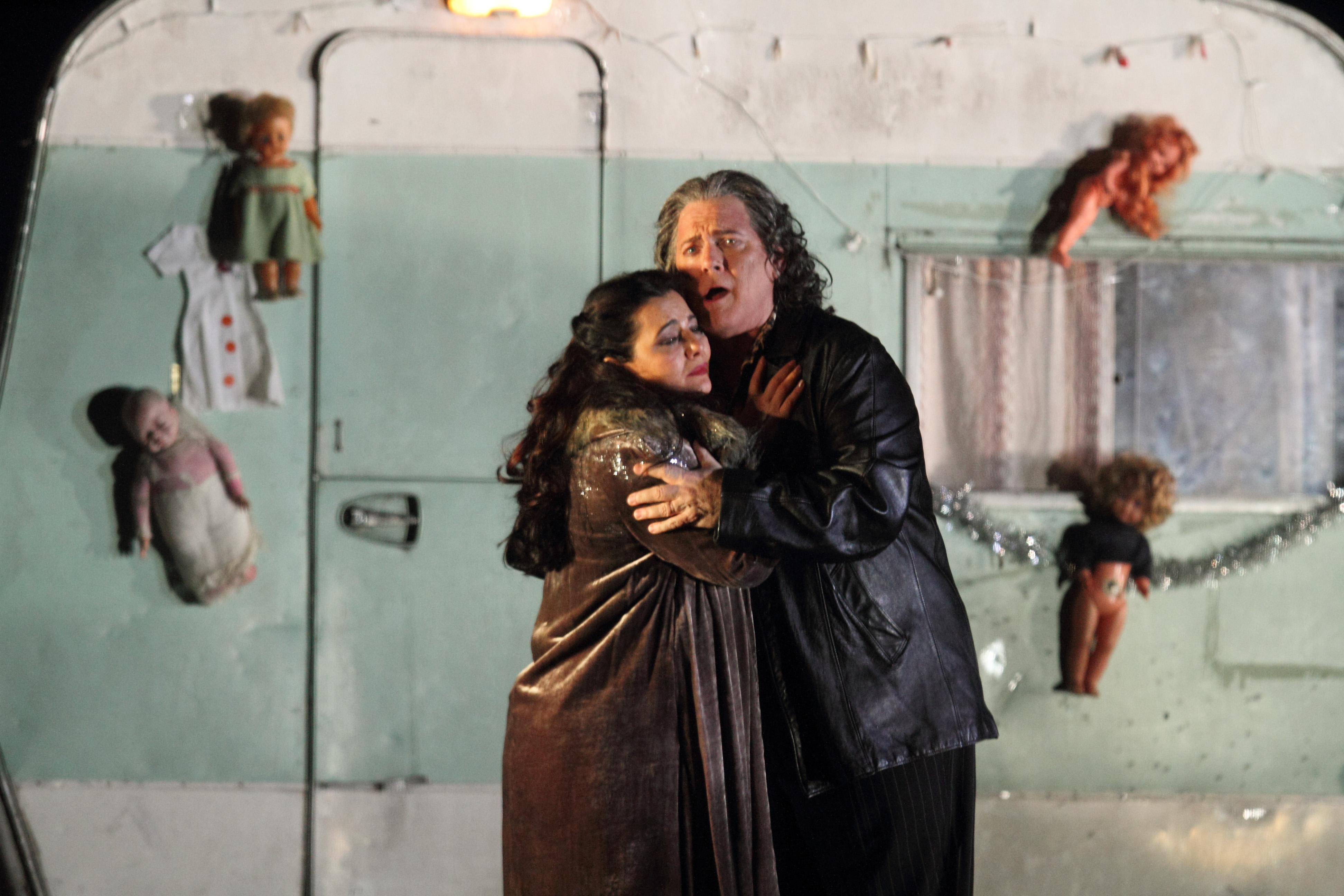Gregory Kunde and  Lianna Haroutounian embrace in front of a blue and white caravan. They both wear leather jackets. Plastic dolls are stuck to the exterior of the caravan behind them.