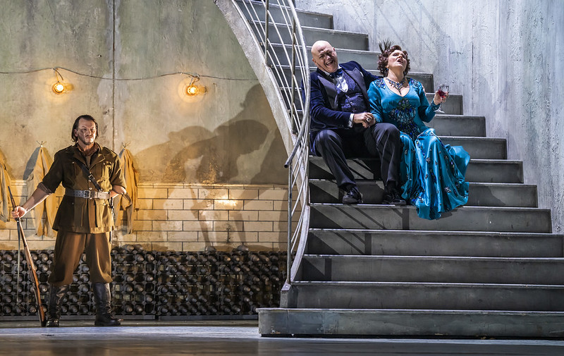  A man in a military uniform stands by a staircase. On the stairs sit a man in a tuxedo and a woman in along blue gown. 