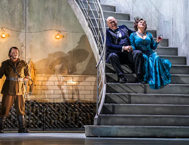 A man in a military uniform stands by a staircase. On the stairs sit a man in a tuxedo and a woman in along blue gown.