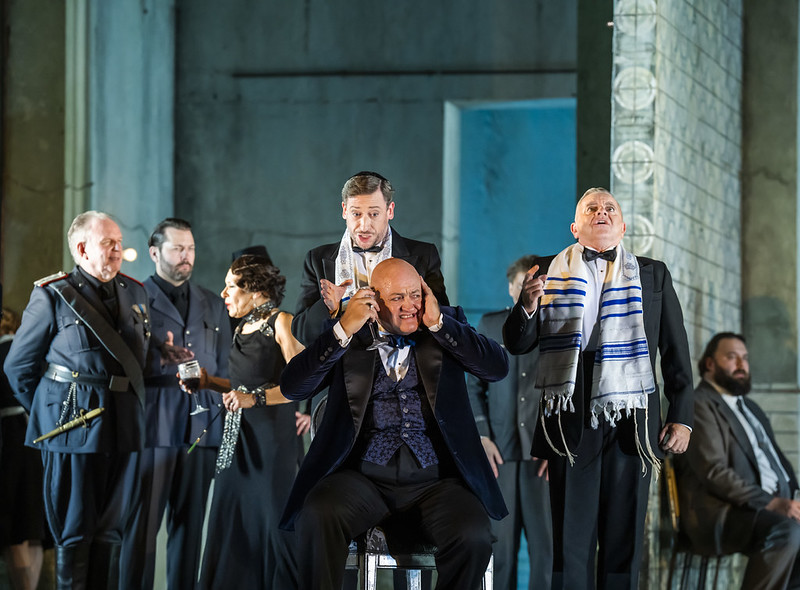 A group of men in black suits and military uniform. One man sits at the center with his hands over his ears dramatically while others stand around him.