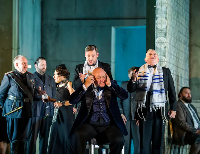 A group of men in black suits and military uniform. One man sits at the center with his hands over his ears dramatically while others stand around him.