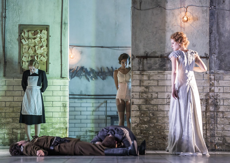 	 A woman in a long white dress stands over two people lying on the floor; a maid and another woman in lingerie stand in the background of an industrial, dimly lit room with tiled walls and exposed light bulbs.