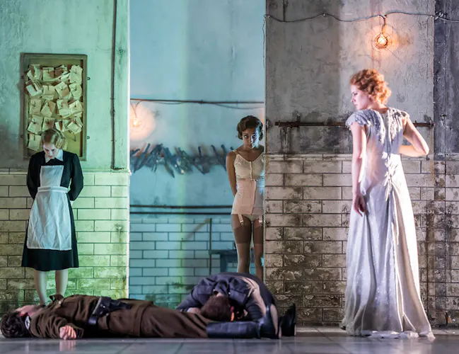 A woman in a long white dress stands over two people lying on the floor; a maid and another woman in lingerie stand in the background of an industrial, dimly lit room with tiled walls and exposed light bulbs.