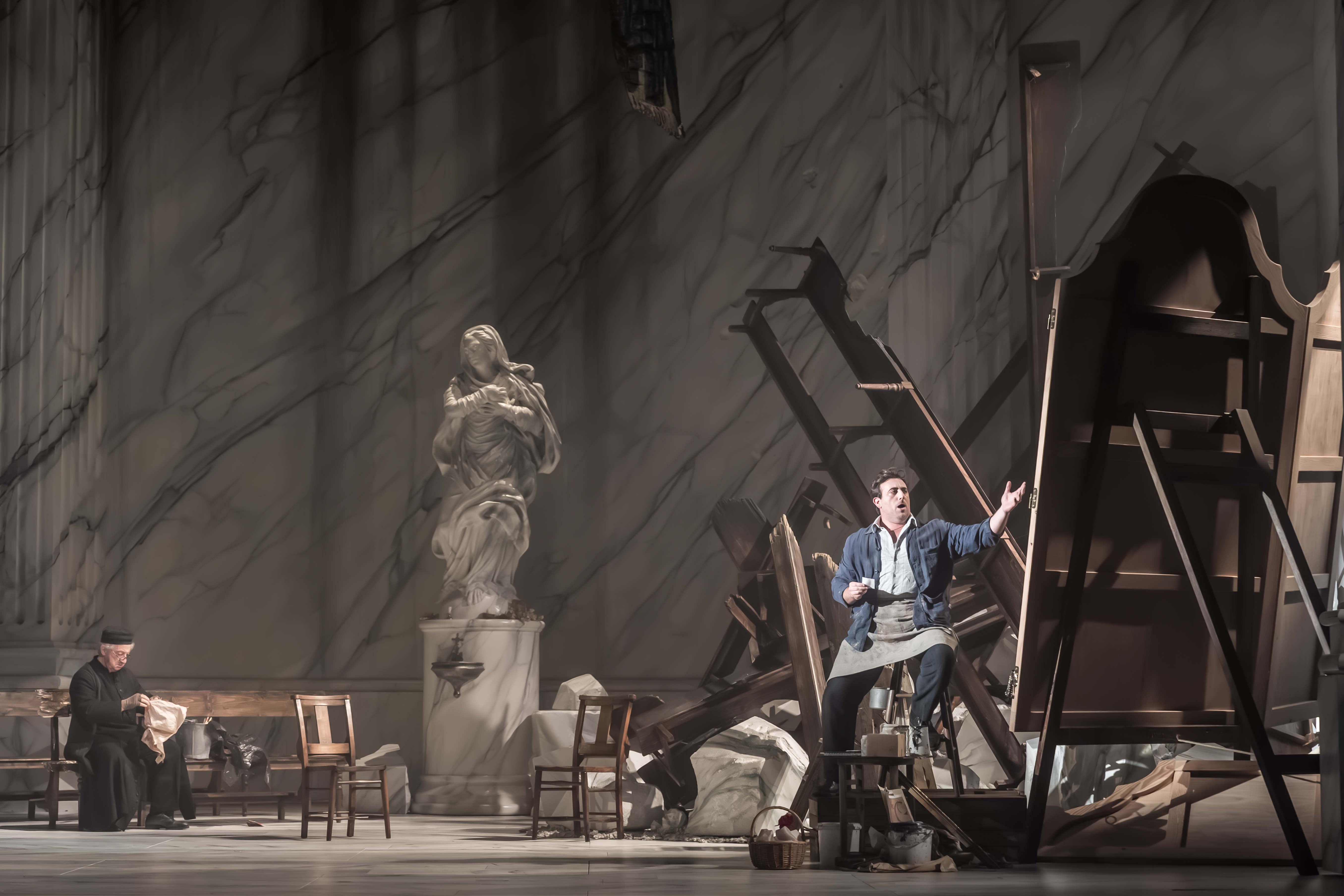 A man gesturing passionately amid overturned furniture and debris, while another person sits reading at left. A large marble statue stands in the background, suggesting a grand, classical setting.