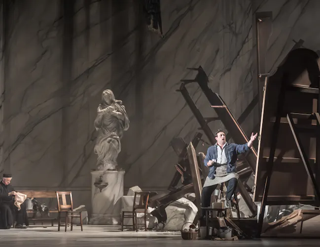 A man gesturing passionately amid overturned furniture and debris, while another person sits reading at left. A large marble statue stands in the background, suggesting a grand, classical setting.