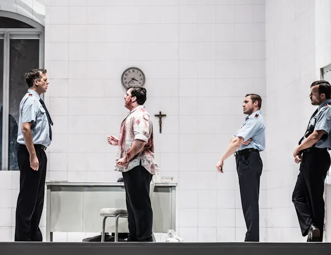 Four men in a stark white room; one in a bloodstained shirt faces three men in police uniforms. A table, chair, clock, and cross are on the wall.