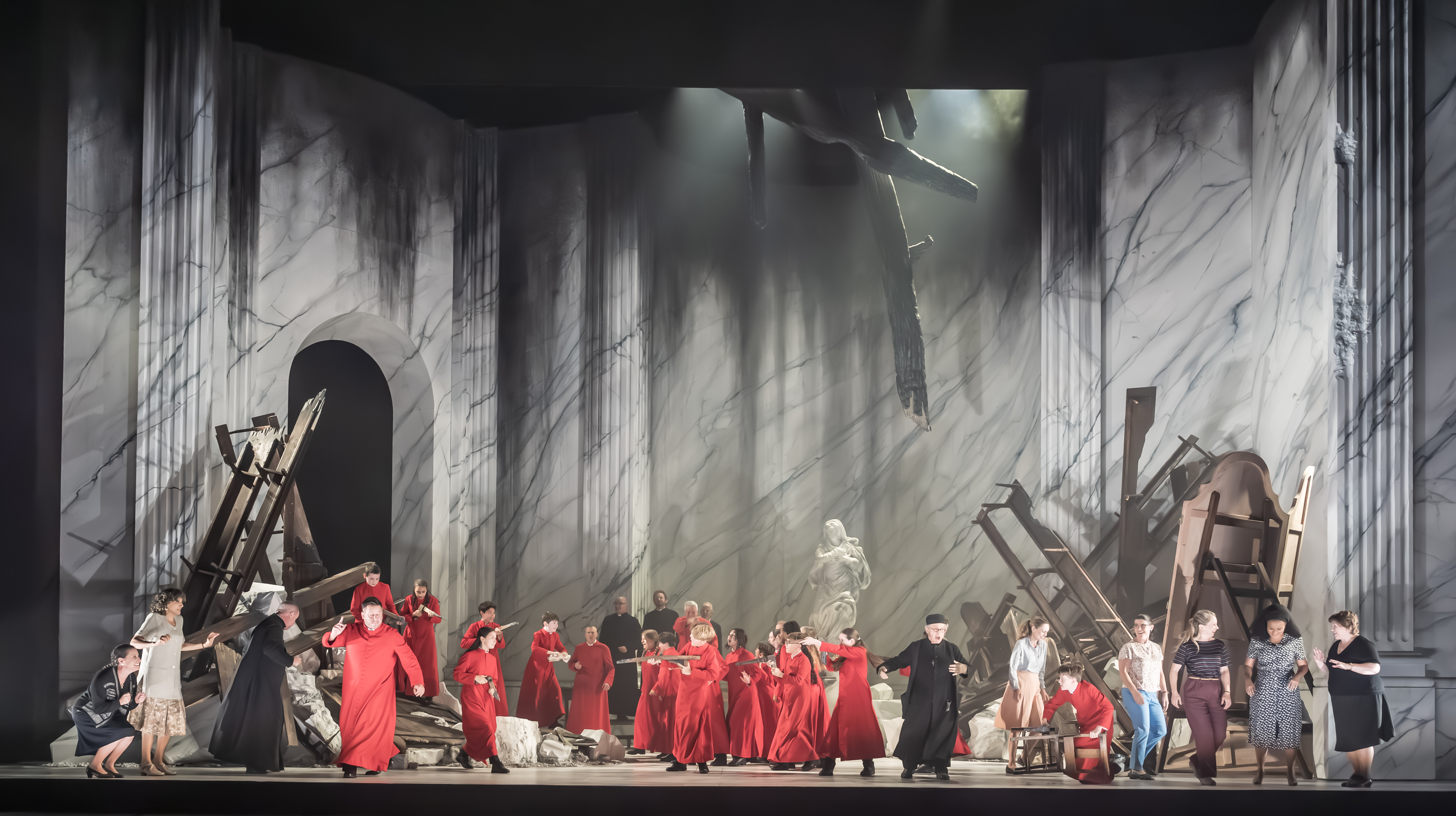 Men in red and black costumes, surrounded by broken columns, rubble, and toppled classical statues, set against a large, marble-patterned backdrop suggestive of ruins.