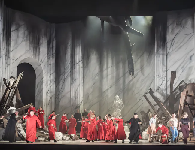 Men in red and black costumes, surrounded by broken columns, rubble, and toppled classical statues, set against a large, marble-patterned backdrop suggestive of ruins.